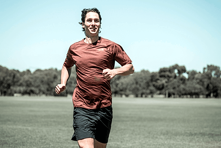 A man in black shorts and a burgundy top is running across an oval with a blue sky above him