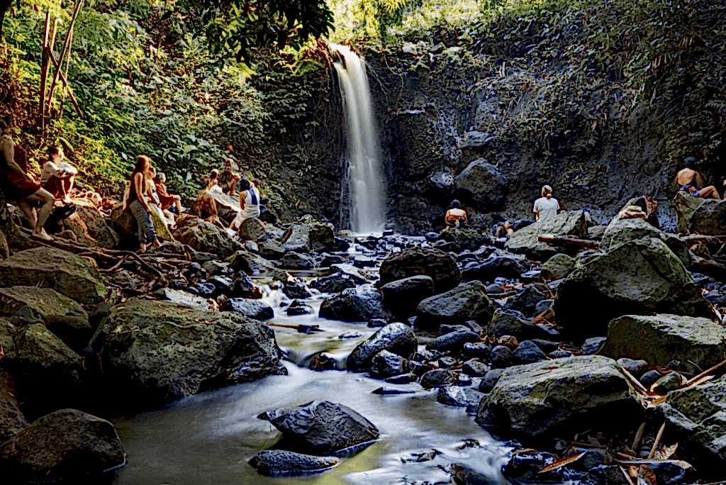 A waterfall rolls down into rockpools. There are lush green trees to the left of the waterfall, and about 10 people are dotted around the frame, sitting on the rocks gazing at the waterfall