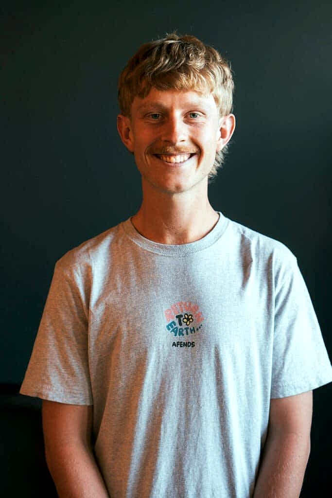A 25 year old man with strawberry blonde hair and a pale blue t-shirt smiles at the camera. He is standing against a dark wall.