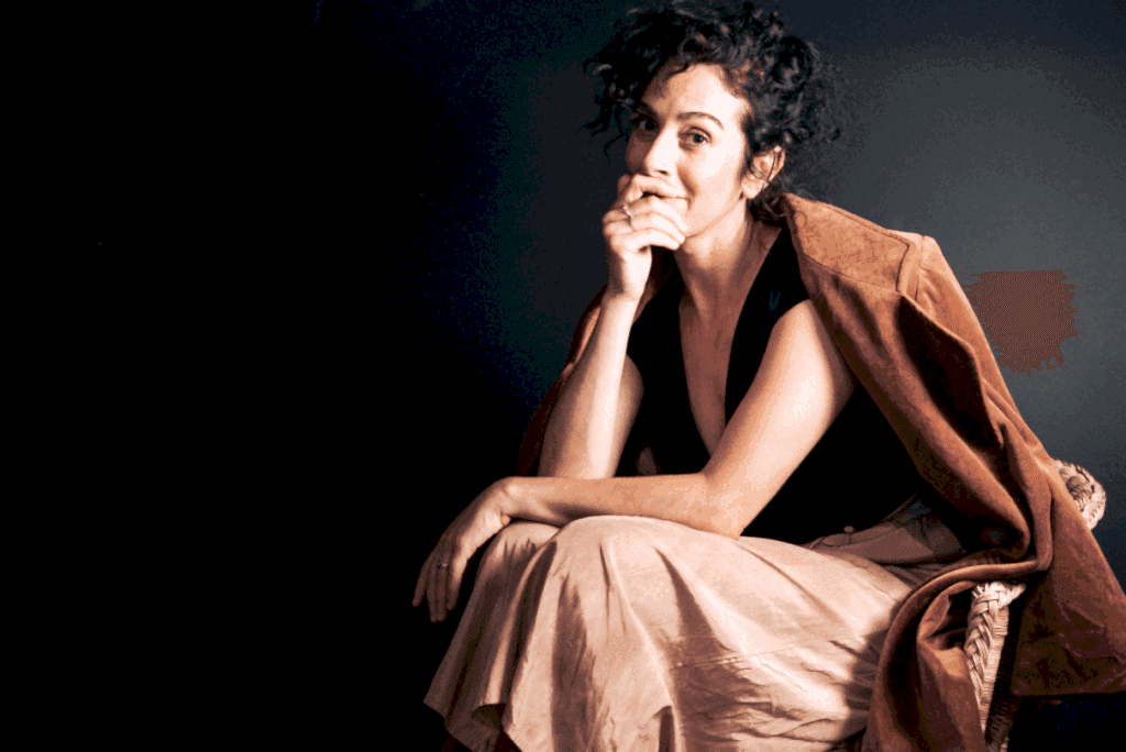 A woman with curly brown hair sits on a chair, facing side on and looking at the camera. She is in a relaxed and contemplative pose with one hand to her face, and wears a beige silky shirt with an orange blazer draped over a black top.