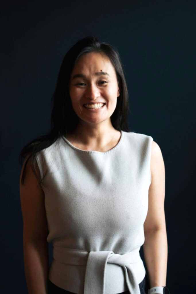 A dark haired woman smiles at the camera, wearing a grey singlet.
