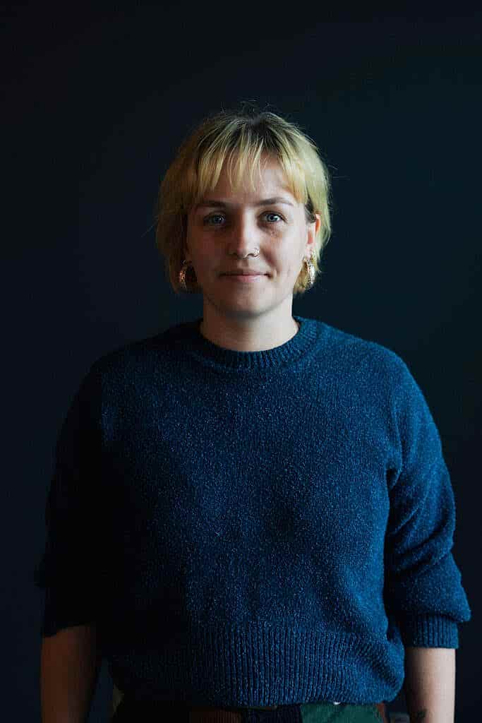 A photo of a woman in a blue sweater. She has a slight smile on her face blonde hair, and is wearing gold earrings.