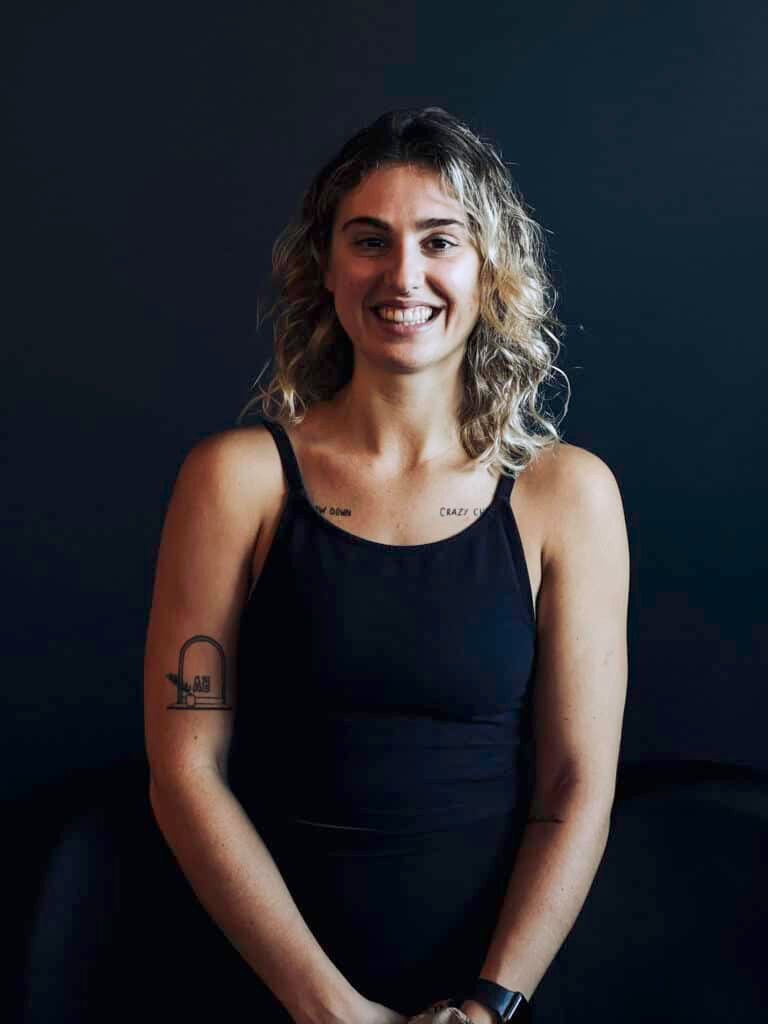 A woman with medium-length blonde curly hair grins at the camera. She is wearing a black singlet and stands against a black background.