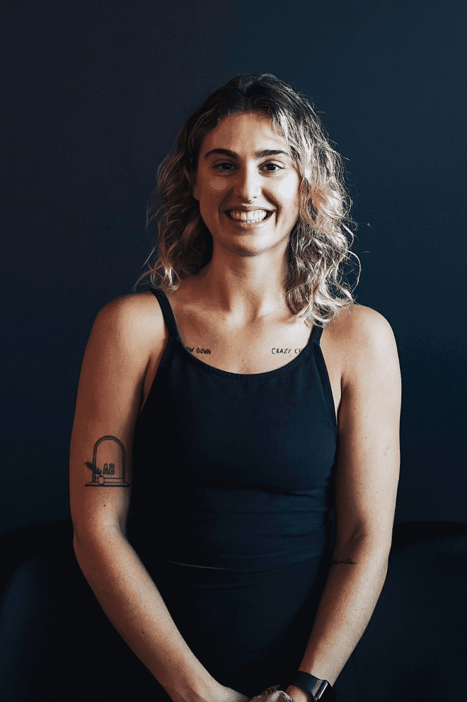 A woman with medium-length blonde curly hair grins at the camera. She is wearing a black singlet and stands against a black background.