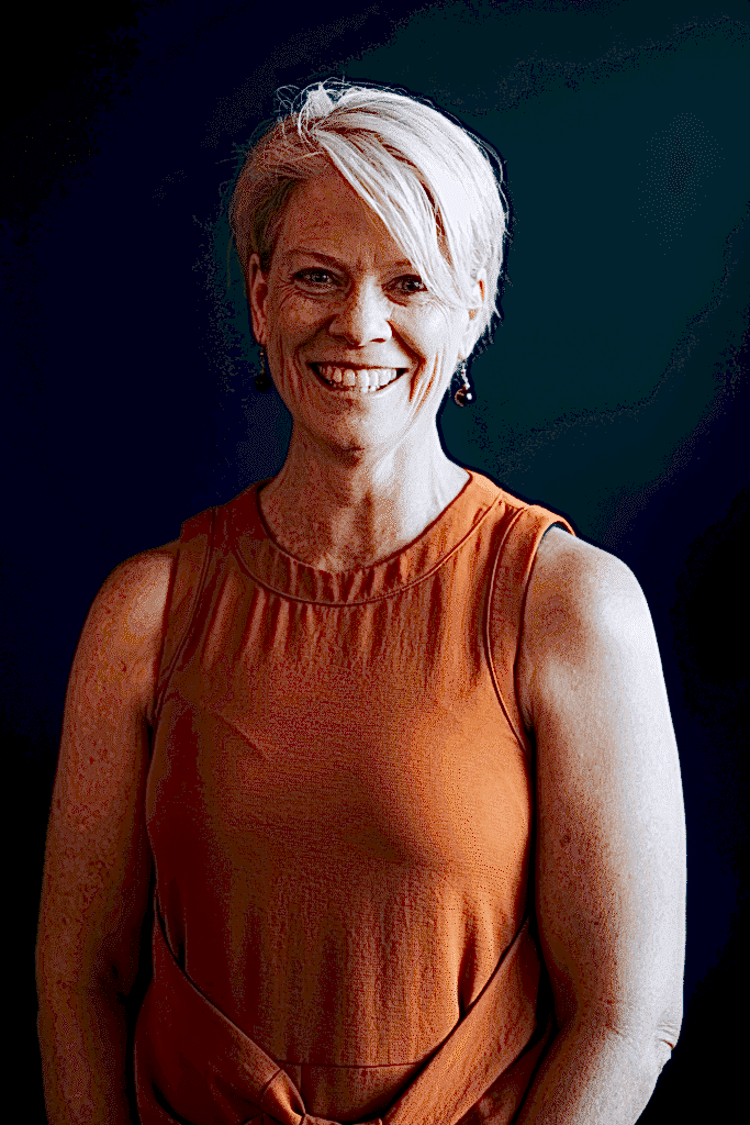 A woman with short, white blonde hair smiles at the camera cheerily. She is against a dark background and wears a burnt orange singlet.