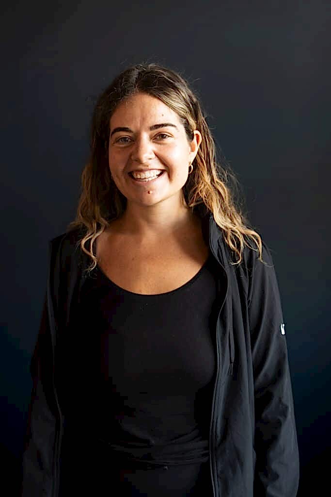 A woman with brown hair smiles in front of a black wall.