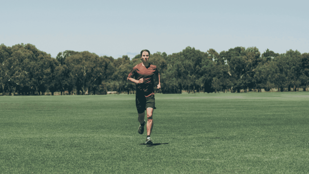 A man runs across a grassy oval in a red shirt and black shorts.