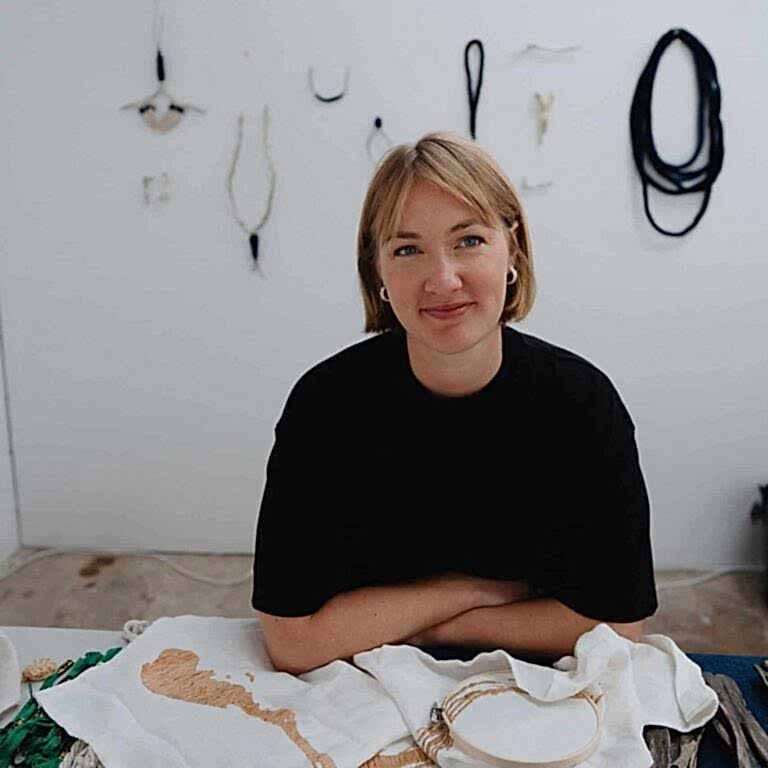 A woman sits at a table working on art with art pieces behind on the wall