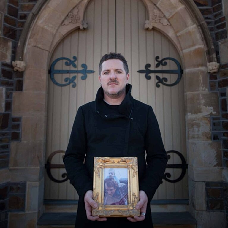 A man stands in front of a church holding a framed family photo